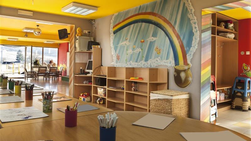 An empty childcare room with a large rainbow painted on the wall, yellow ceilings and a table full of coloring pencils and markers