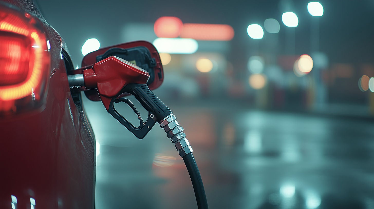 Red car refueling at night in the rain, gas pump nozzle inserted in fuel tank, city lights blurred in background.