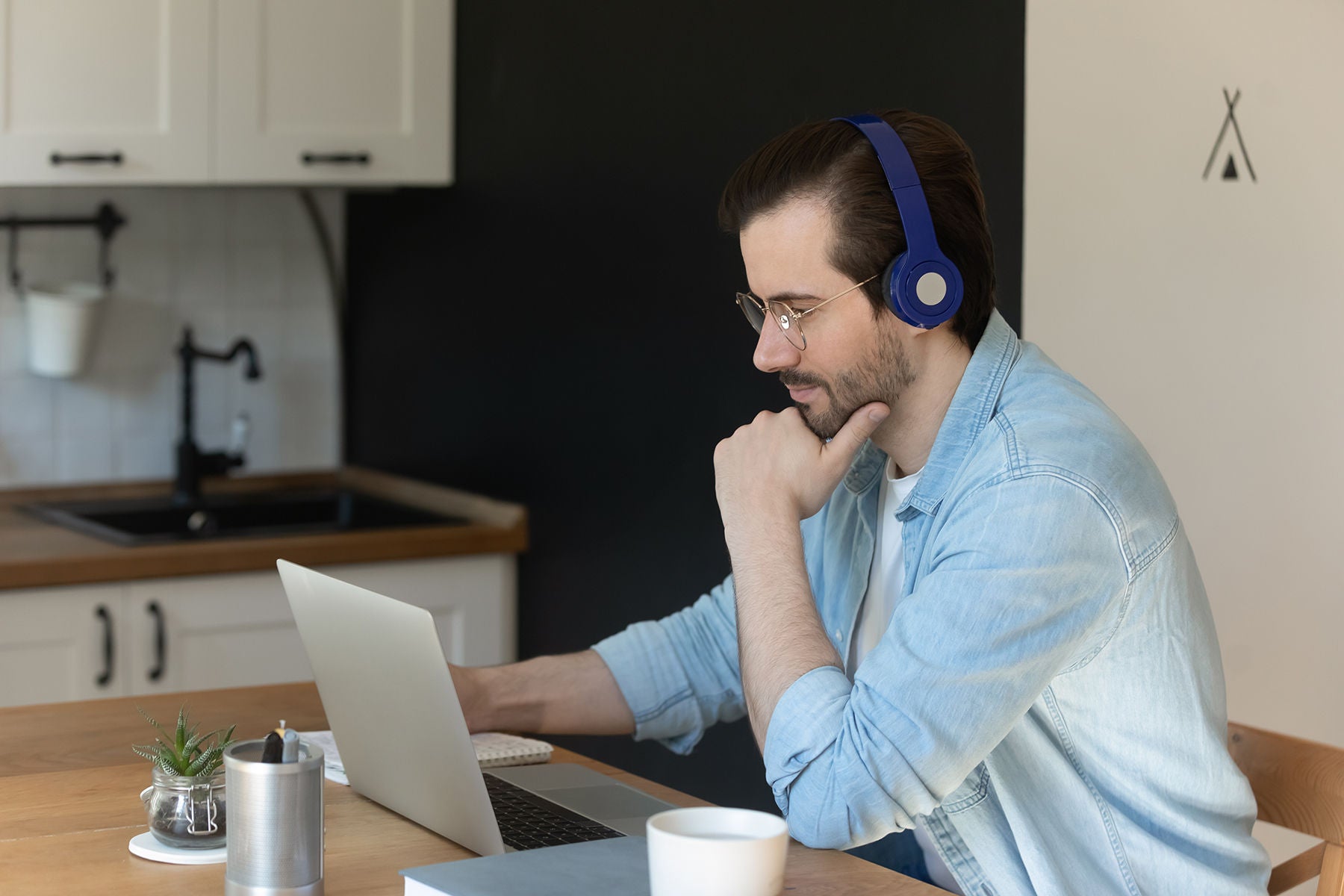 Man in wireless headphones look at laptop screen while sitting at kitchen table