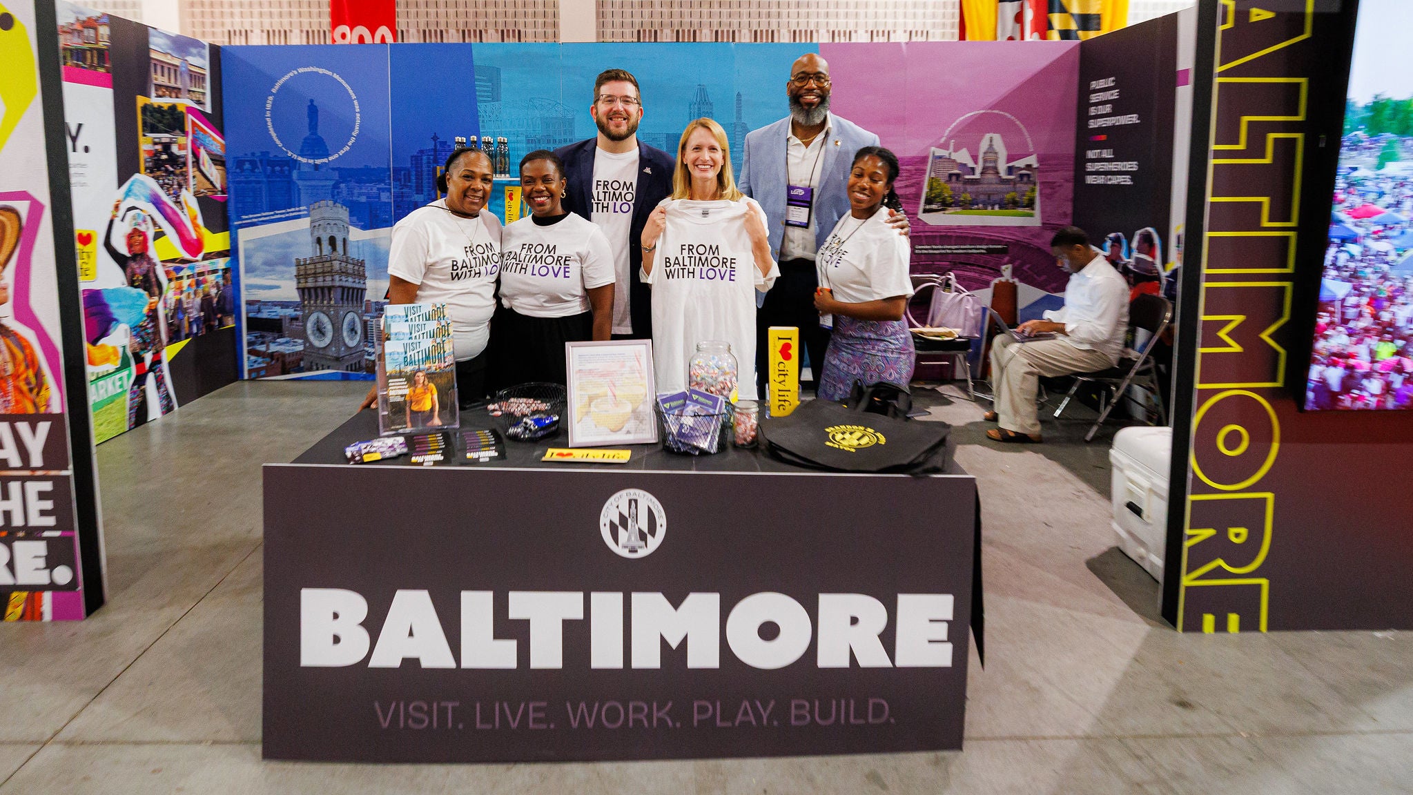 Comptroller Lierman standing with a group of people at a table that says Baltimore, Visit, Live, Work , Play, Build. She is holding up a T-shirt that says From Baltimore with Love