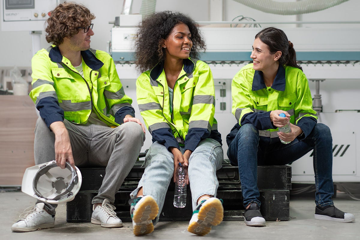 Break time. Group of diverse engineer people sitting together take a break and talking. Young multiracial woman and colleagues technician relax after job in factory. Manufacturing employee lifestyle.