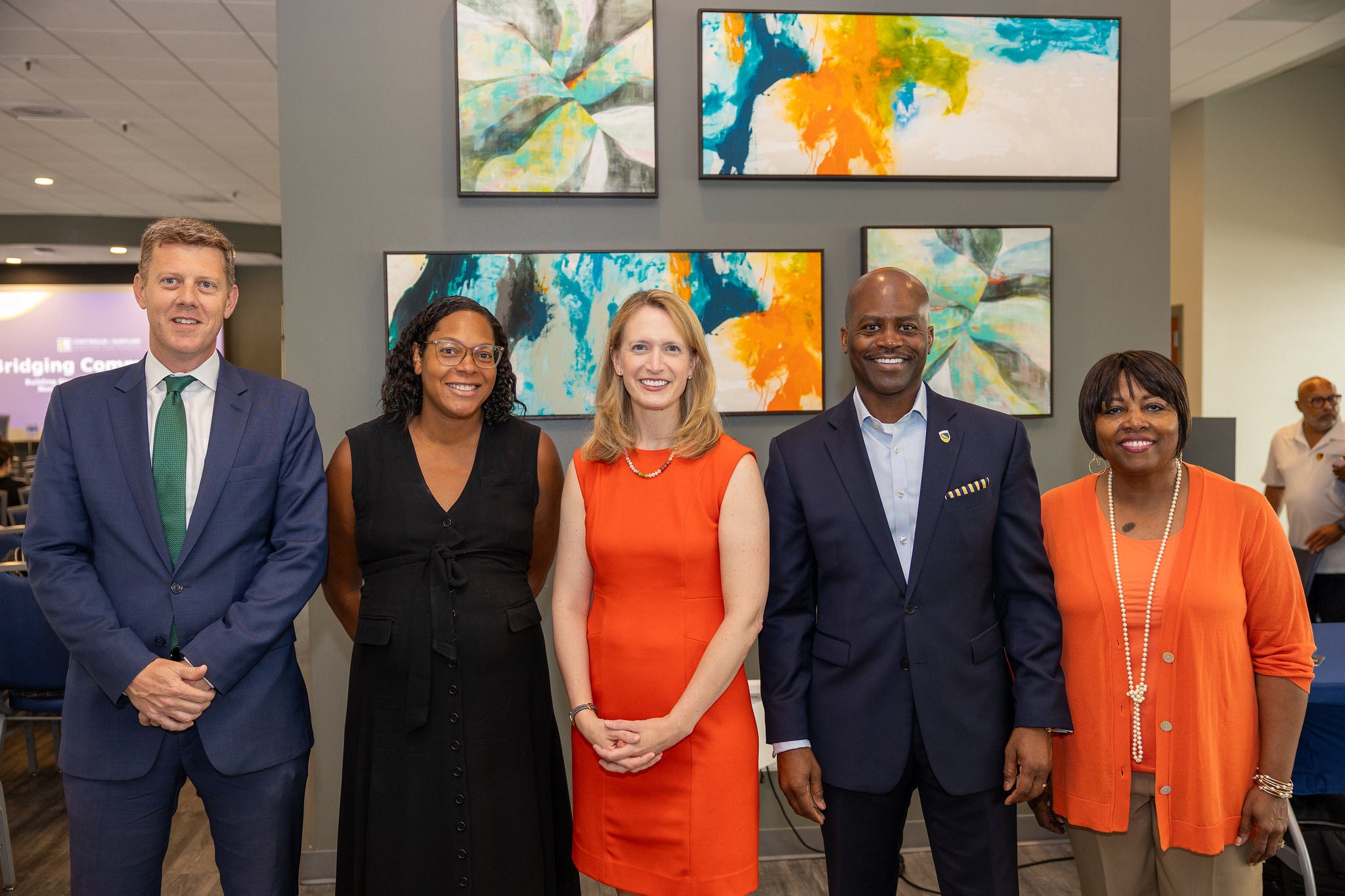 Comptroller Lierman weating an orange dress standing with 4 other people in front of a wall of colorful paintings