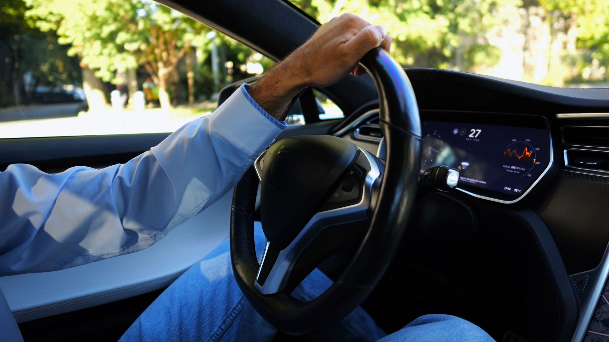 Male hand of businessman holds steering wheel while driving electrical vehicle at city. Businessperson operating electric car on urban road. Young man commuting to work at sunny day.