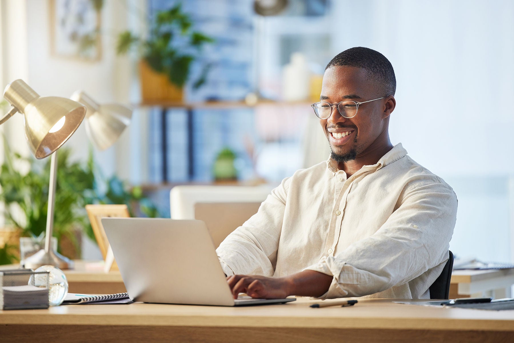 Man with glasses sitting at a desk smiling while typing on a laptop
