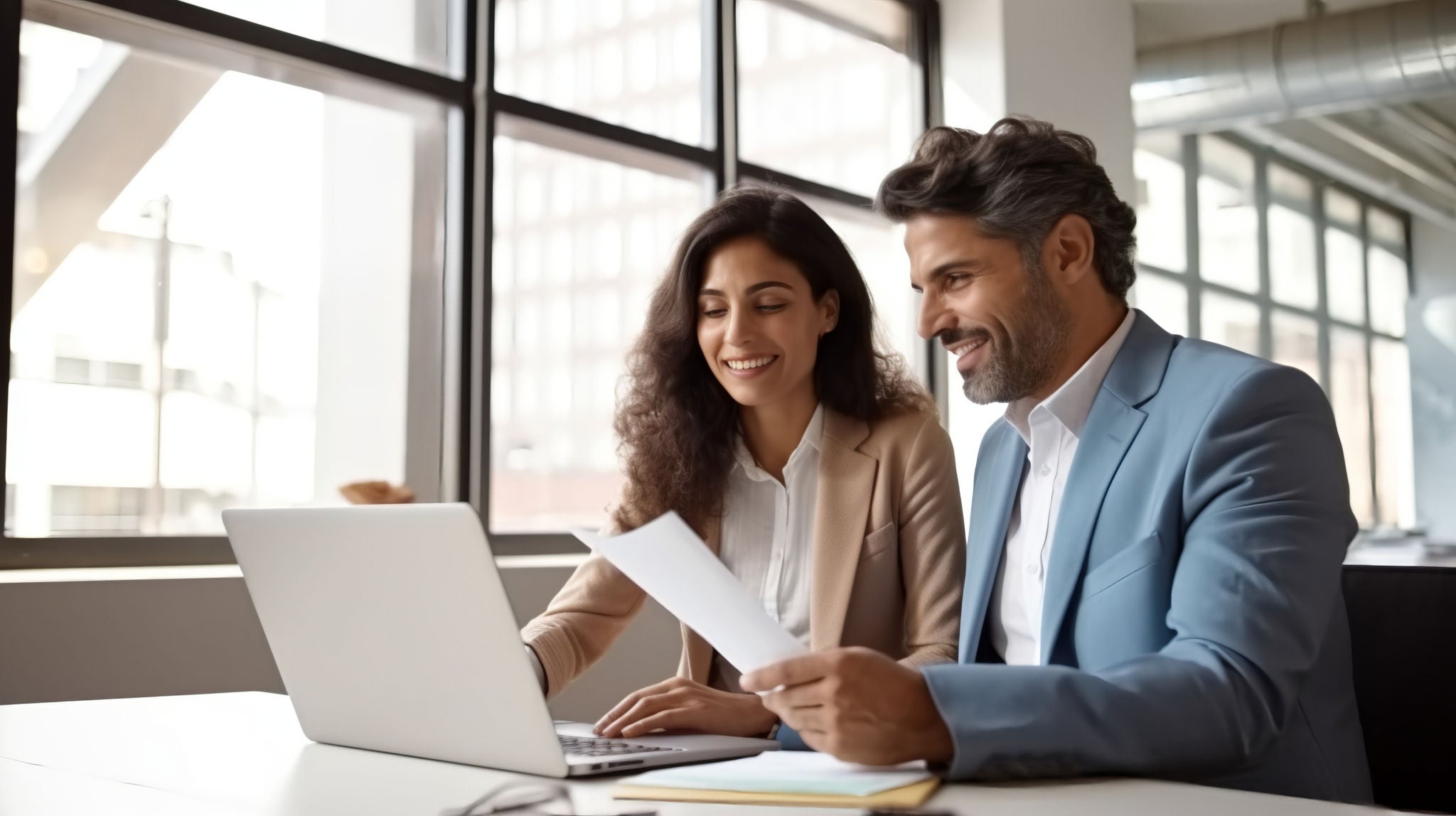 A man and woman dressed smartly, are sitting at an office desk with a laptop open as they look over documents together, smiling.