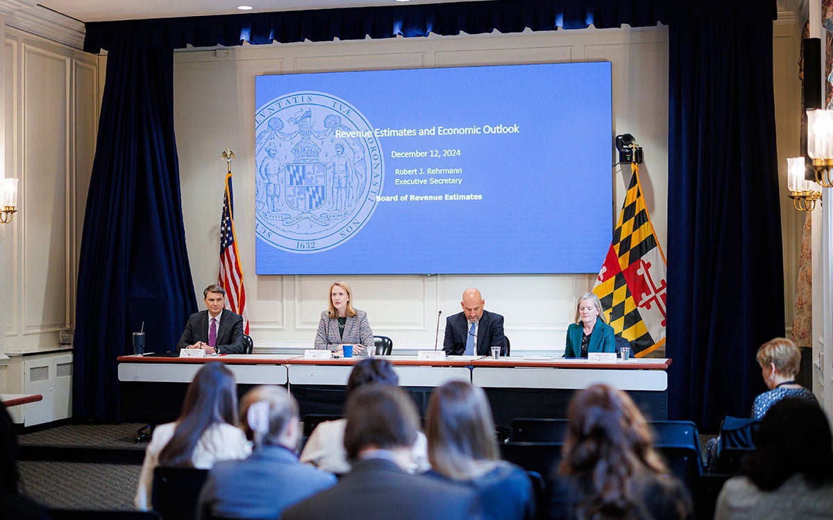 People sitting at a table with a large screen behind them addressing a audience